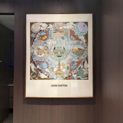 A large square framed art display on a dark wood wall featuring a silk scarf with intricate vintage travel illustrations, airships, trains, and iconic landmarks in pastels.