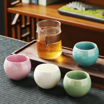 Four pastel-colored spherical ceramic teacups with ice-crackle glazes arranged on a dark tea mat beside a clear textured glass tea pitcher with a wooden accent collar.