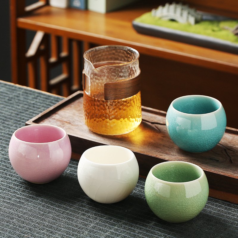 Four pastel-colored spherical ceramic teacups with ice-crackle glazes arranged on a dark tea mat beside a clear textured glass tea pitcher with a wooden accent collar.