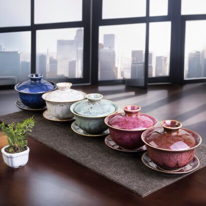 Five ceramic gaiwan tea sets lined up in a row on a dark table runner in front of a city skyline window, featuring distinct ice-crackle glaze colors including cobalt blue, off-white, celadon green, magenta pink, and amber brown.