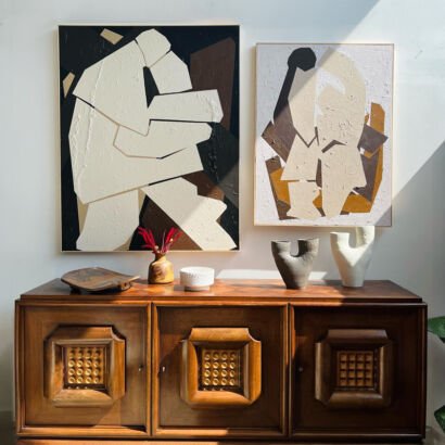 Two large framed abstract canvas paintings featuring bold geometric collage shapes in beige, black, brown, and ochre tones hanging on a white wall above a vintage wooden sideboard.