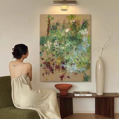 A large vertical canvas art piece with expressive green, white, and burgundy abstract floral brushstrokes hanging on a beige wall under a gallery light, next to a tall white vase and a person sitting on an armchair.