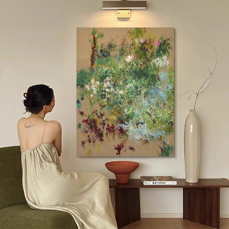 A large vertical canvas art piece with expressive green, white, and burgundy abstract floral brushstrokes hanging on a beige wall under a gallery light, next to a tall white vase and a person sitting on an armchair.