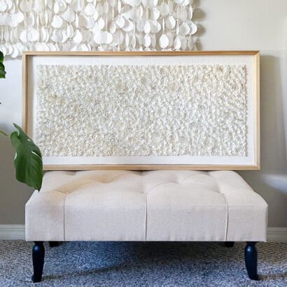 A large horizontal framed canvas artwork with dense, impasto white floral rosette textures resting on a beige tufted ottoman with a light wood frame.