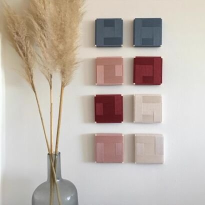 Eight small square woven yarn panels arranged in a grid on a white wall, featuring muted blue, blush pink, deep burgundy, and cream geometric patterns beside pampas grass.