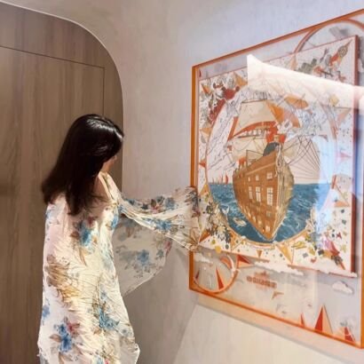 A large square acrylic floating frame with an orange border displaying a fine silk scarf artwork featuring a grand nautical sailing ship on open sea, mounted on a neutral wall.