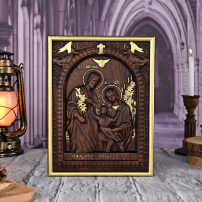 An upright rectangular wooden wall plaque featuring a detailed high-relief carving of the Holy Family with golden accents and Cyrillic inscriptions, enclosed within a gold-trimmed carved frame, displayed on a wooden surface between a vintage lantern and a lit candle.