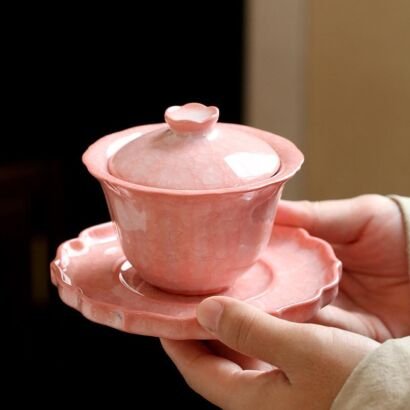 A pastel pink handcrafted ceramic gaiwan lidded teacup with subtle ice-crackle glaze textures resting on a matching flower-rim saucer held in two hands.