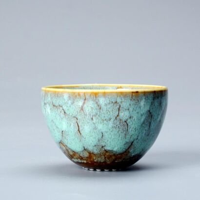 A small round ceramic tea bowl featuring a light turquoise reactive glaze with rustic brown crackle veins and an earthy dark base set against a plain gray background.