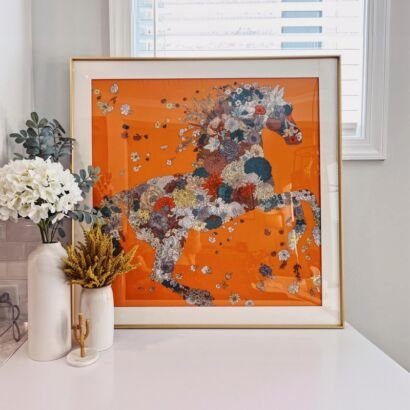 A square luxury framed silk scarf wall art featuring a rearing horse made of intricate floral patterns on a vivid orange background, set in a gold frame.