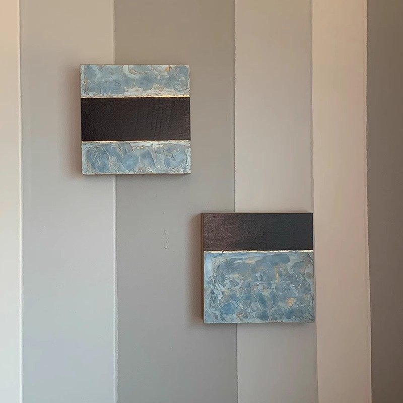 Two square abstract canvas paintings featuring thick-textured light blue and dark charcoal color blocking separated by thin metallic accents, mounted on a neutral striped wall.