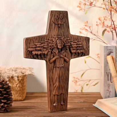 An upright cross-shaped wooden wall plaque featuring a detailed high-relief carving of Jesus as the Good Shepherd holding a lamb amidst a flock of sheep, with a dove symbol above, displayed on a wooden surface next to a pinecone, basket, and book.