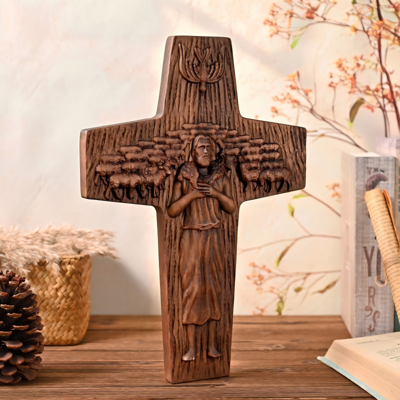 An upright cross-shaped wooden wall plaque featuring a detailed high-relief carving of Jesus as the Good Shepherd holding a lamb amidst a flock of sheep, with a dove symbol above, displayed on a wooden surface next to a pinecone, basket, and book.