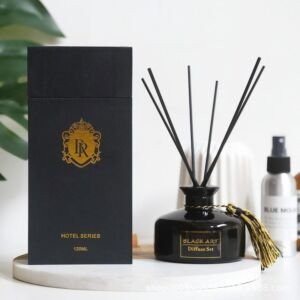 A sleek black glass reed diffuser bottle with black sticks and a gold tassel accent, displayed next to its matte black gift box with gold foil emblem stamping.