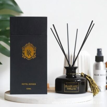 A sleek black glass reed diffuser bottle with black sticks and a gold tassel accent, displayed next to its matte black gift box with gold foil emblem stamping.