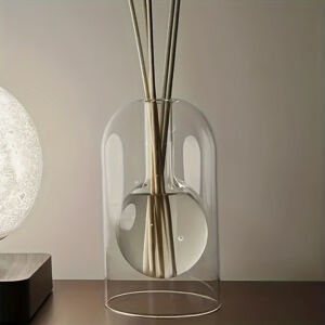 A sleek double-walled clear glass reed diffuser bottle featuring an inner spherical bulb holding essential oil and diffuser sticks, set beside a soft glowing table lamp.
