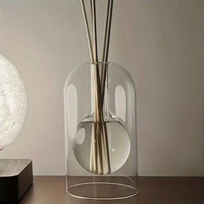 A sleek double-walled clear glass reed diffuser bottle featuring an inner spherical bulb holding essential oil and diffuser sticks, set beside a soft glowing table lamp.