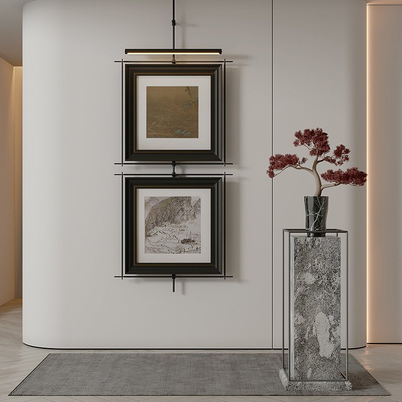 Two vertically stacked framed Asian landscape artworks hanging from a black metal rod with an integrated picture light next to a potted red bonsai tree on a marble pedestal.