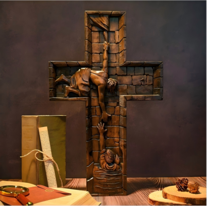 An upright cross-shaped wooden wall plaque featuring a detailed high-relief carving of a figure reaching down from within a brick-textured structure to rescue another figure emerging from water, displayed on a wooden surface next to books and pinecones.