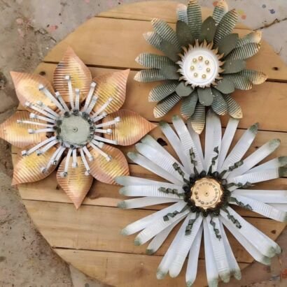 Three rustic layered metal flower wall art set, textured metal floral hanging decor in golden, sage green and off-white finishes displayed on wooden plank board