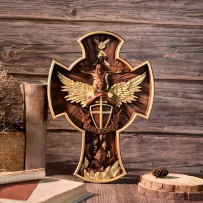 An upright cross-shaped wooden wall plaque featuring a detailed high-relief carving of Saint Michael with golden wings, a shield, and the inscription "SAINT MICHAEL", along with other sacred figures, enclosed within a gold-accented carved wooden cross frame, displayed on a wooden surface.