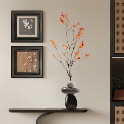 Stacked stone sculpture vase in smooth light gray, charcoal, and black holding tall delicate branches with orange autumn leaves on a dark console table