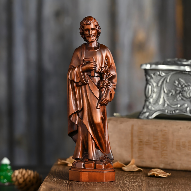 A standing carved dark wood figurine of Saint Joseph holding a carpenter's square and a sprig of lilies, displayed on a wooden surface next to an old book and a decorative silver box.