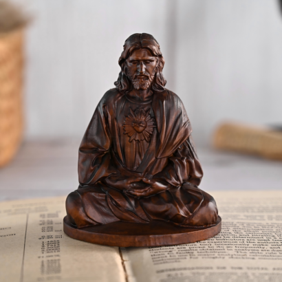 A dark wood carved figurine of Jesus sitting in a meditative cross-legged pose with hands clasped in his lap and the Sacred Heart symbol on his chest, resting on an open book.