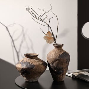Two distressed, earth-toned wabi-sabi ceramic vases with weathered clay textures, one containing a bare branch ornament, resting on a dark table surface