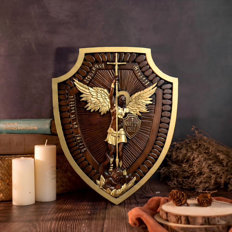 An upright shield-shaped wooden wall plaque featuring a detailed high-relief carving of Archangel Michael with golden wings and accents subduing a dragon, displayed on a table next to candles and pinecones.