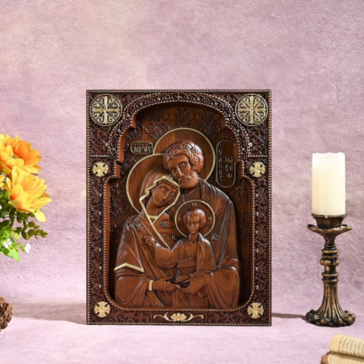 An upright rectangular wooden icon plaque featuring a detailed high-relief carving of the Holy Family with golden lettering and accents within an intricately carved border, displayed on a table next to yellow flowers and a lit candle on a holder.