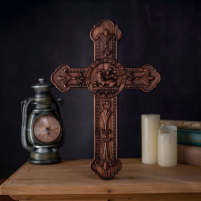 An upright wooden cross plaque featuring intricate relief carvings and a central circular medallion depicting the Agnus Dei (Lamb of God) with a banner, displayed on a wooden surface next to a vintage lantern, candles, and books.
