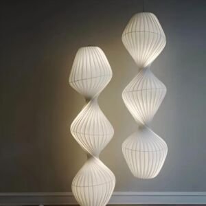 A modern sculptural floor lamp and matching hanging pendant light with white pleated fabric twisted in a three-tier spiral silhouette emitting a warm soft glow against a grey wall background.