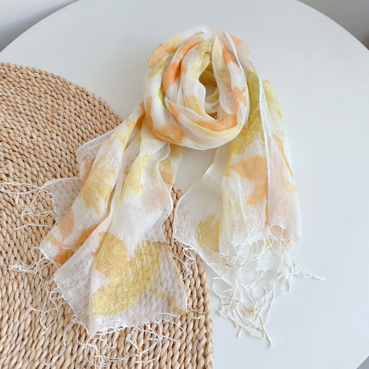 A lightweight off-white sheer scarf featuring soft watercolor floral patterns in pastel orange and yellow shades, finished with twisted tassel fringe edges and draped on a round woven straw placemat over a white table.