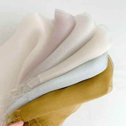A layered fan display of sheer organza silk scarves in soft pastel shades including dusty pink, off-white, pale blue, light grey, and mustard yellow with delicate fringe edges on a white background.