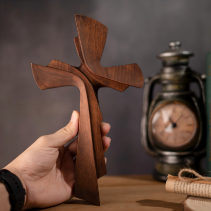 A hand holding a smooth, modern sculptural wooden cross with flowing, overlapping curved lines and a rich dark wood finish, displayed against a blurred background with a clock and rolled paper.