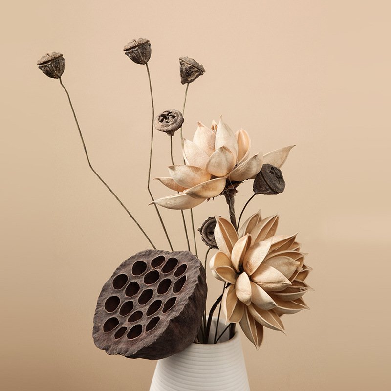 Minimalist dried floral arrangement featuring natural dark lotus pods, poppy seed heads, and carved wood flowers in a white ceramic vase against a beige background