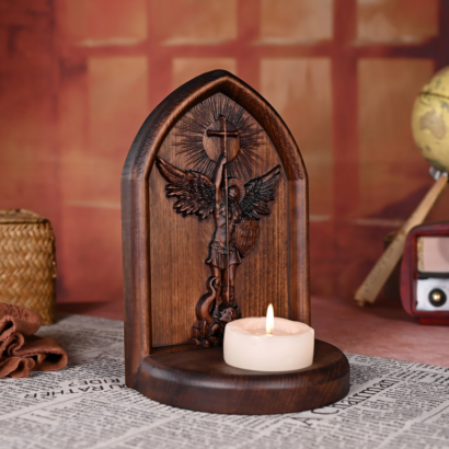 An upright arch-shaped wooden plaque featuring a detailed high-relief carving of Archangel Michael subduing a dragon with a cross-topped staff, with a built-in circular base holding a lit white tea candle, displayed on a newspaper-patterned surface.