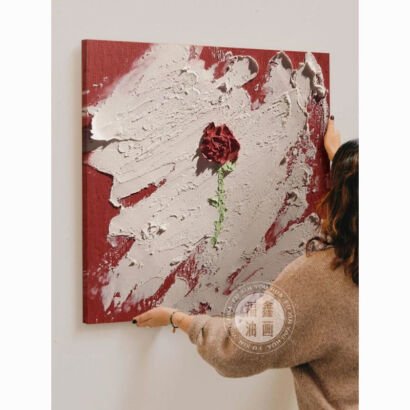 A woman hanging a square 3D textured canvas painting featuring a sculpted red rose centered on a plaster-style white and deep red impasto background.