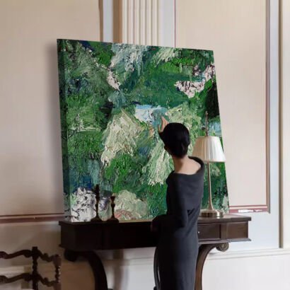 A woman in a dark dress admiring a large, heavily textured green and white impasto abstract painting resting on a vintage wooden console table next to a lamp.