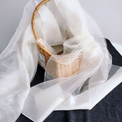 A lightweight off-white sheer organza scarf artfully draped over and flowing out of a natural woven wicker basket on a dark contrasting background.