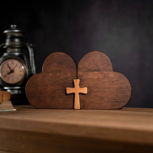 Dark wood double heart sculpture joined together by a light wood cross sitting on a wooden surface next to a vintage clock lantern