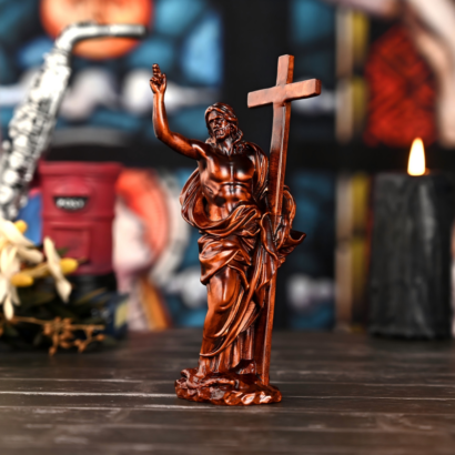 A dark wood carved figurine of Jesus Christ standing upright and holding a large cross with one hand while raising the other hand in blessing, displayed on a table against a blurred background with a lit candle and decorative objects.
