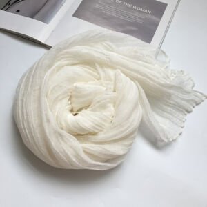 A lightweight off-white sheer scarf with a crinkled, pleated texture artfully swirled into a rose-like spiral next to an open magazine on a white surface.