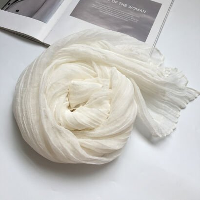 A lightweight off-white sheer scarf with a crinkled, pleated texture artfully swirled into a rose-like spiral next to an open magazine on a white surface.