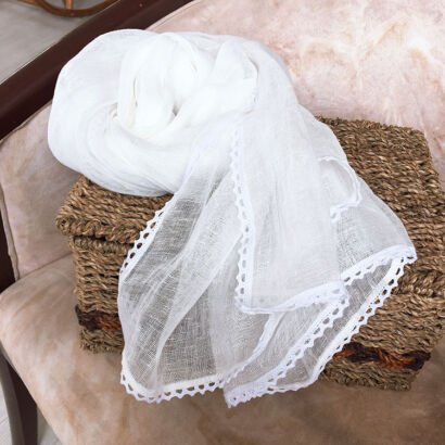 A pure white, lightweight semi-sheer cotton-linen scarf featuring delicate crocheted lace edges, elegantly swirled over a woven straw storage basket on a plush beige chair.