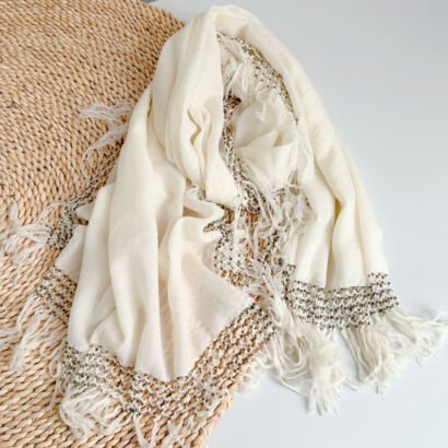An off-white or ivory lightweight linen-cotton scarf featuring a woven mesh crochet openwork border with dark contrast stitches and long frayed fringe tassels, laid out over a woven straw placemat.