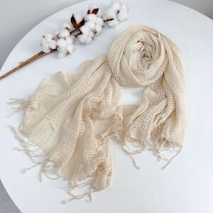 A cream-colored or off-white open-weave mesh knit scarf with lightweight breathable texture and knotted fringe tassels, artfully swirled on a white surface next to a decorative dried cotton stalk.
