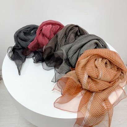 A display of thin, lightweight sheer scarves with wavy horizontal stripes arranged in a row on a white table, available in various colors including orange, olive green, taupe brown, dark red, and black.