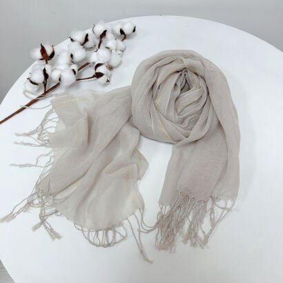 A light beige or off-white lightweight linen scarf with subtle woven gold metallic lurex stripes and knotted fringe ends, neatly swirled on a white surface alongside a decorative cotton stalk.